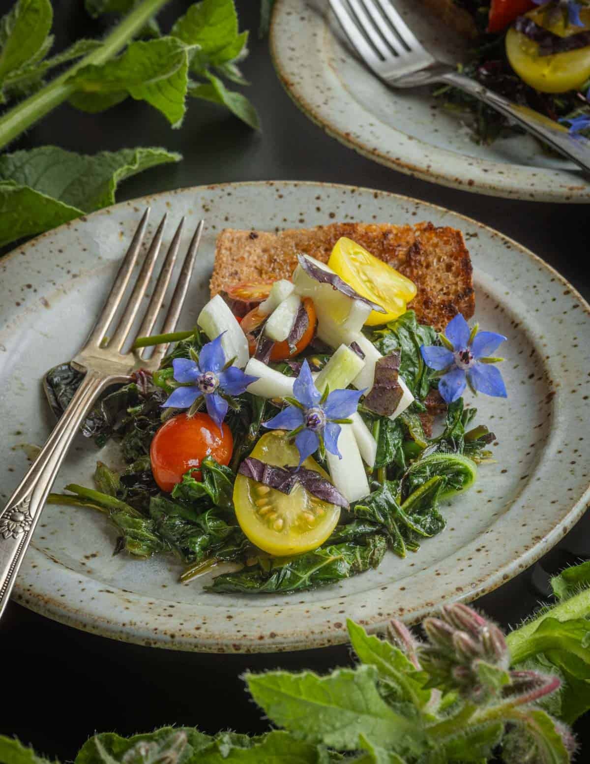 Cooked borage, comfrey, and beet greens on a plate garnished with fresh tomatoes, cucumbers and borage flowers.