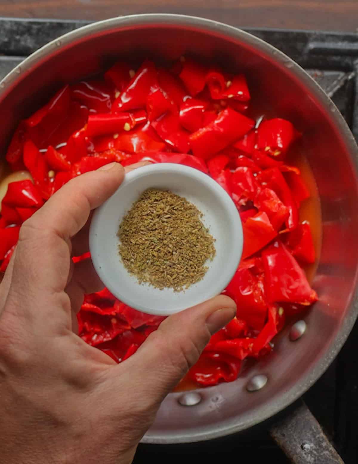A hand holding a small dish showing ground cumin to be added to a pan of hot sauce.