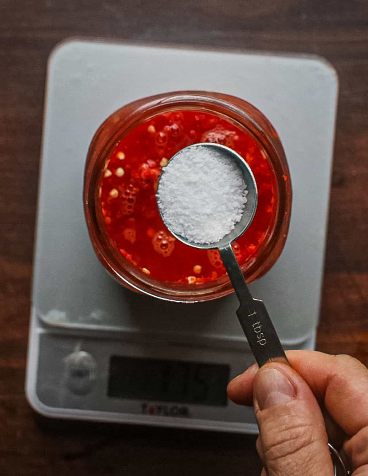 A measuring spoon filled with salt hovering over a jar of ground Fresno peppers and water on a scale.