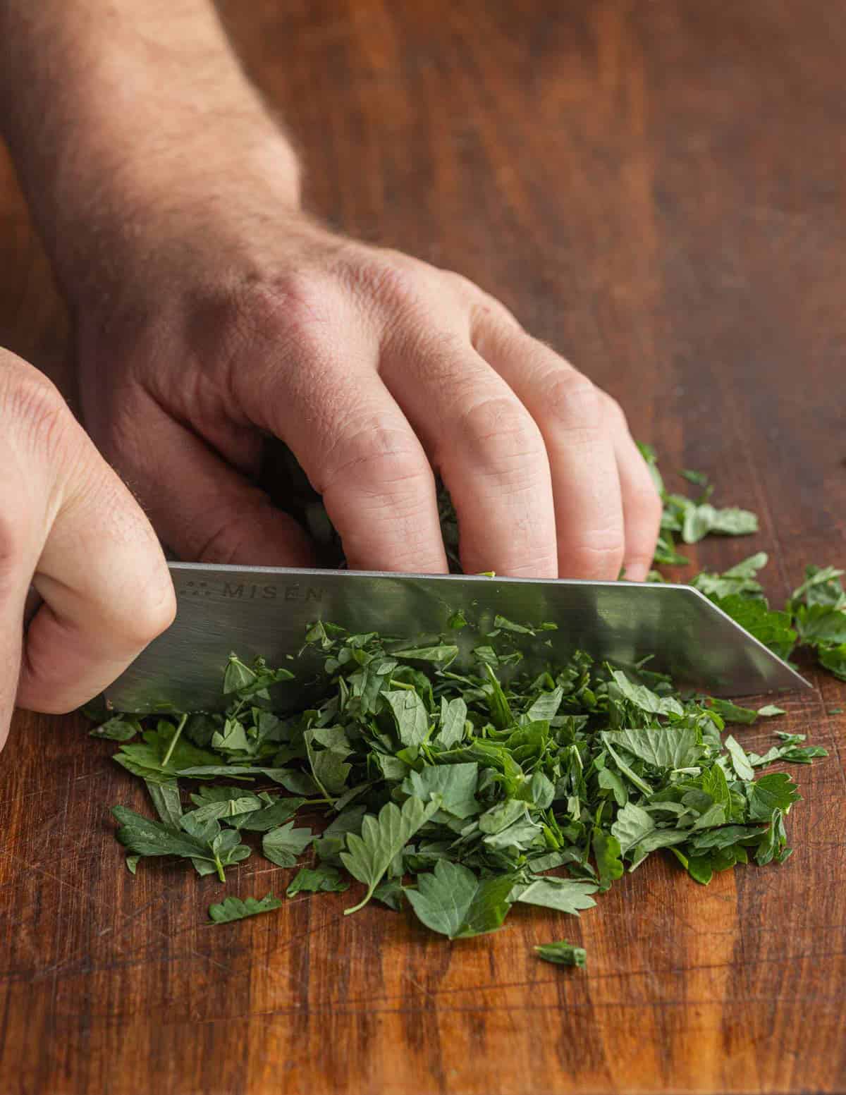 A hand with a knife finely chopping salad burnet leaves on a cutting board.