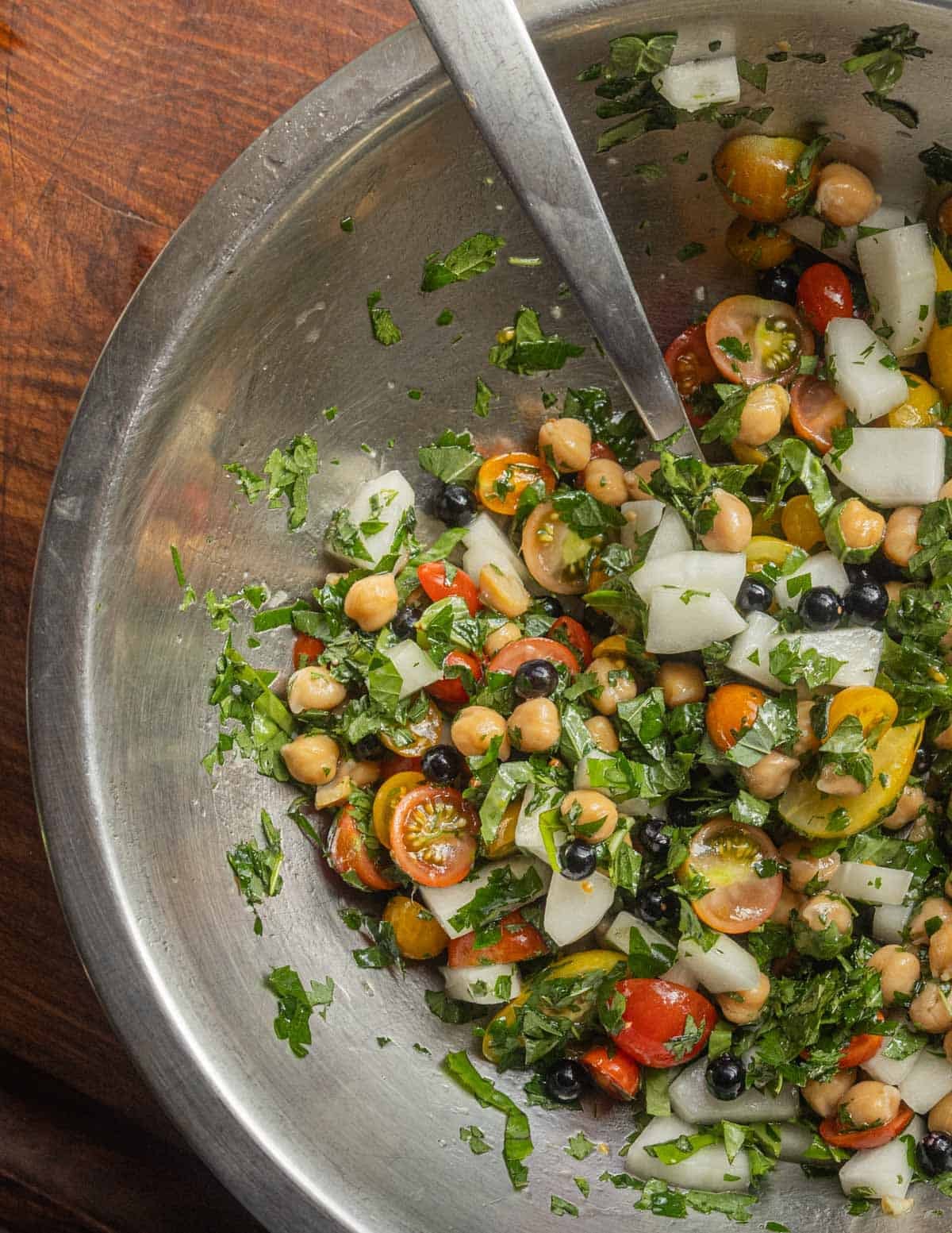 Mixing cucumbers, tomatoes, and chickpeas with herbs and chopped salad burnet leaves in a bowl.