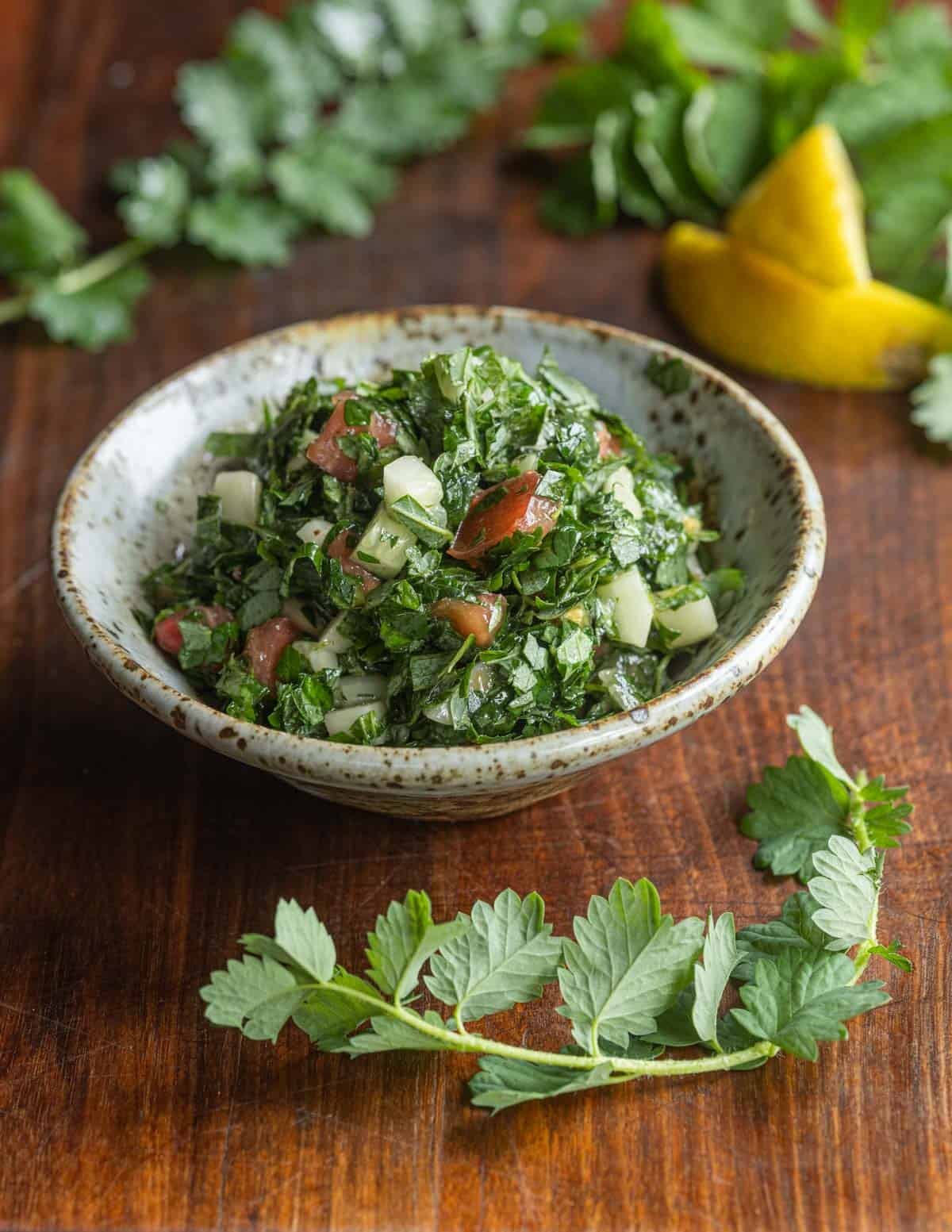 A bowl of tabouleh made with minced salad burnet leaves instead of parsley on a wood background with fresh sprigs of salad burnet around the bowl.