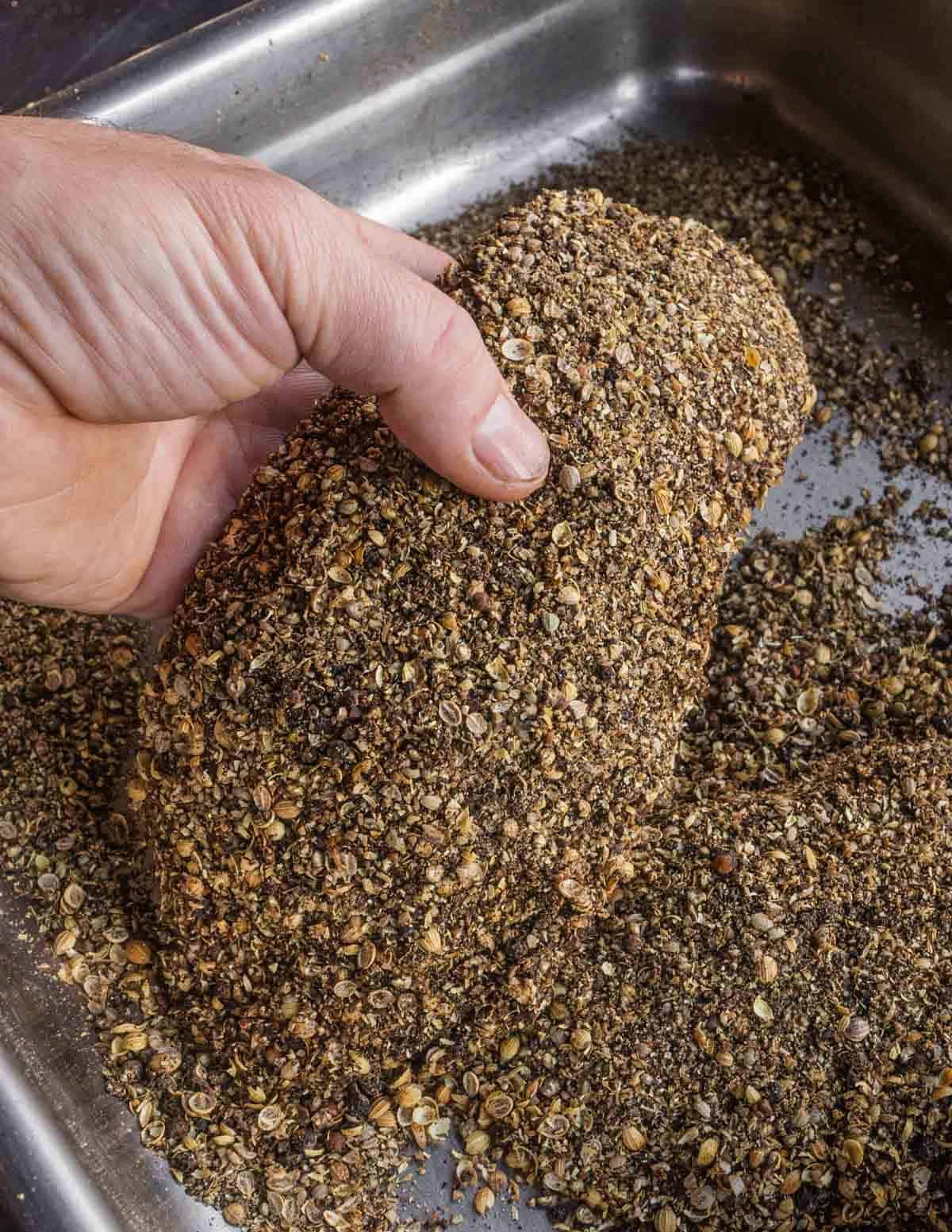A hand holding a cured goose breast crusted with peppercorns and coriander.