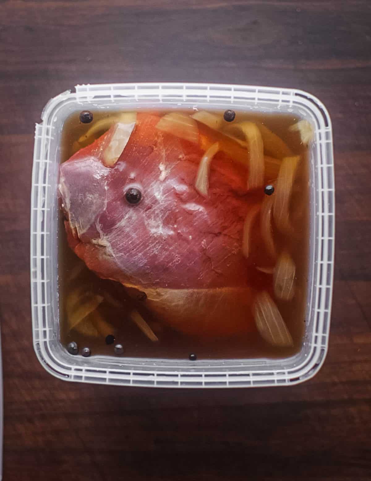 A goose breast immersed in a pastrami brine in a square deli container.