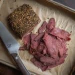 Thinly shaved goose pastrami on a baking sheet lined with parchment paper next to a knife.