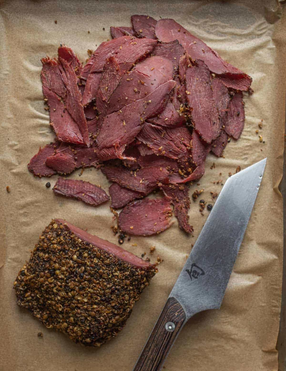 Sliced goose pastrami on a baking sheet lined with parchment next to a slicing knife.