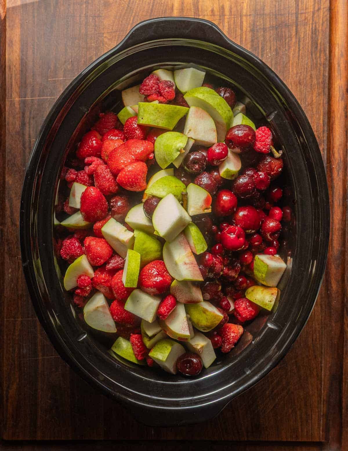 A top down image of a slow-cooker filled with cut up cherries, apples, pears, blueberries and cranberries before cooking.
