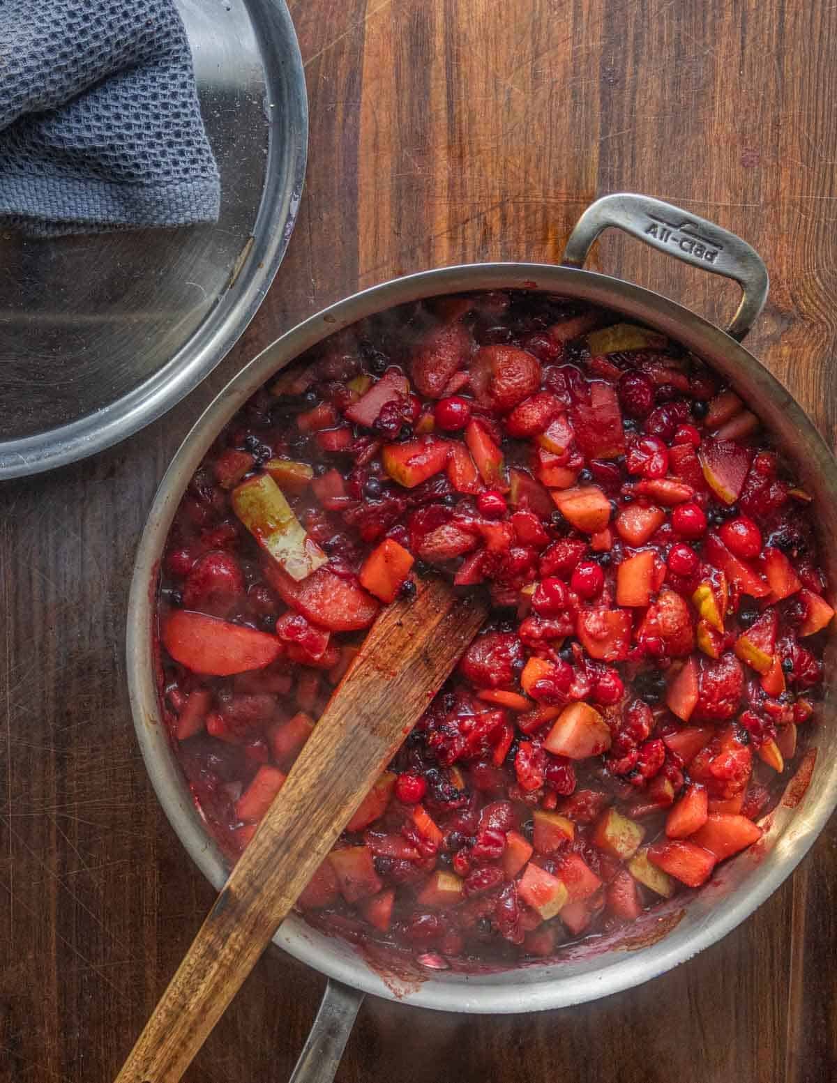 A top down image of an all clad pan filled with cooked apples, cranberries, blueberries and strawberries.