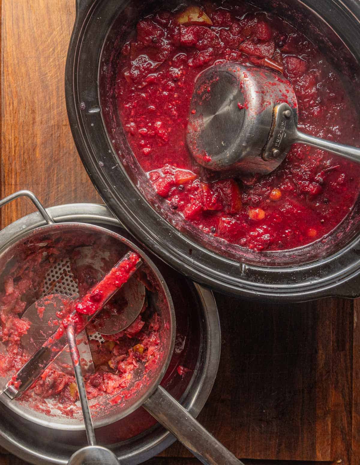 A top down image of a slow cooker filled with cooked fruits being ladled into a food mill and strained.