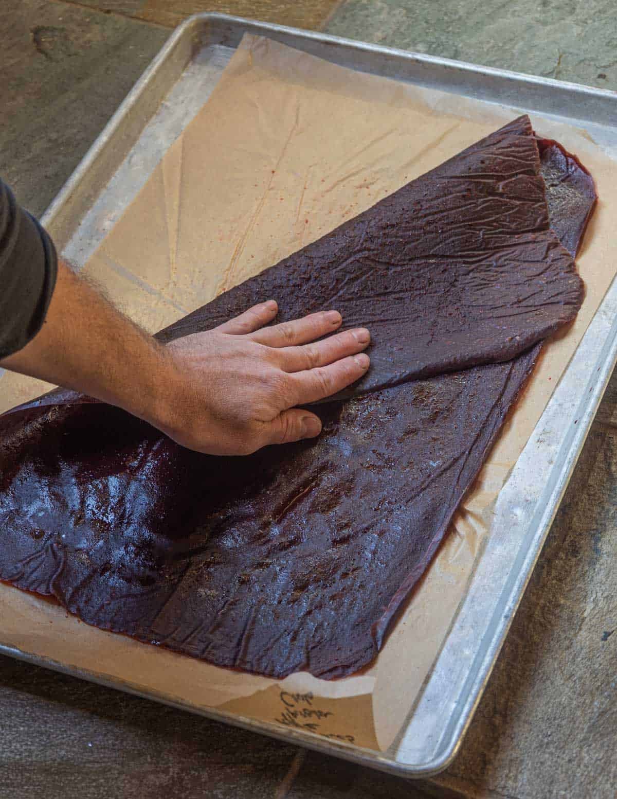 A hand folding over a sheet of fruit leather on a full sized baking sheet lined with parchment.