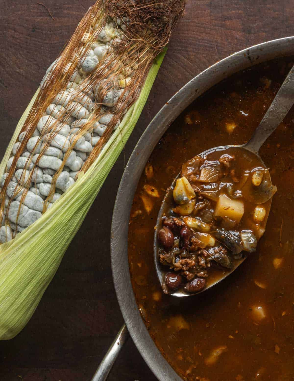 Sopa de huitlaoche con chorizo. Corn mushroom soup with potatoes and black beans in a pot next to an ear of fresh huitlacoche mushrooms.