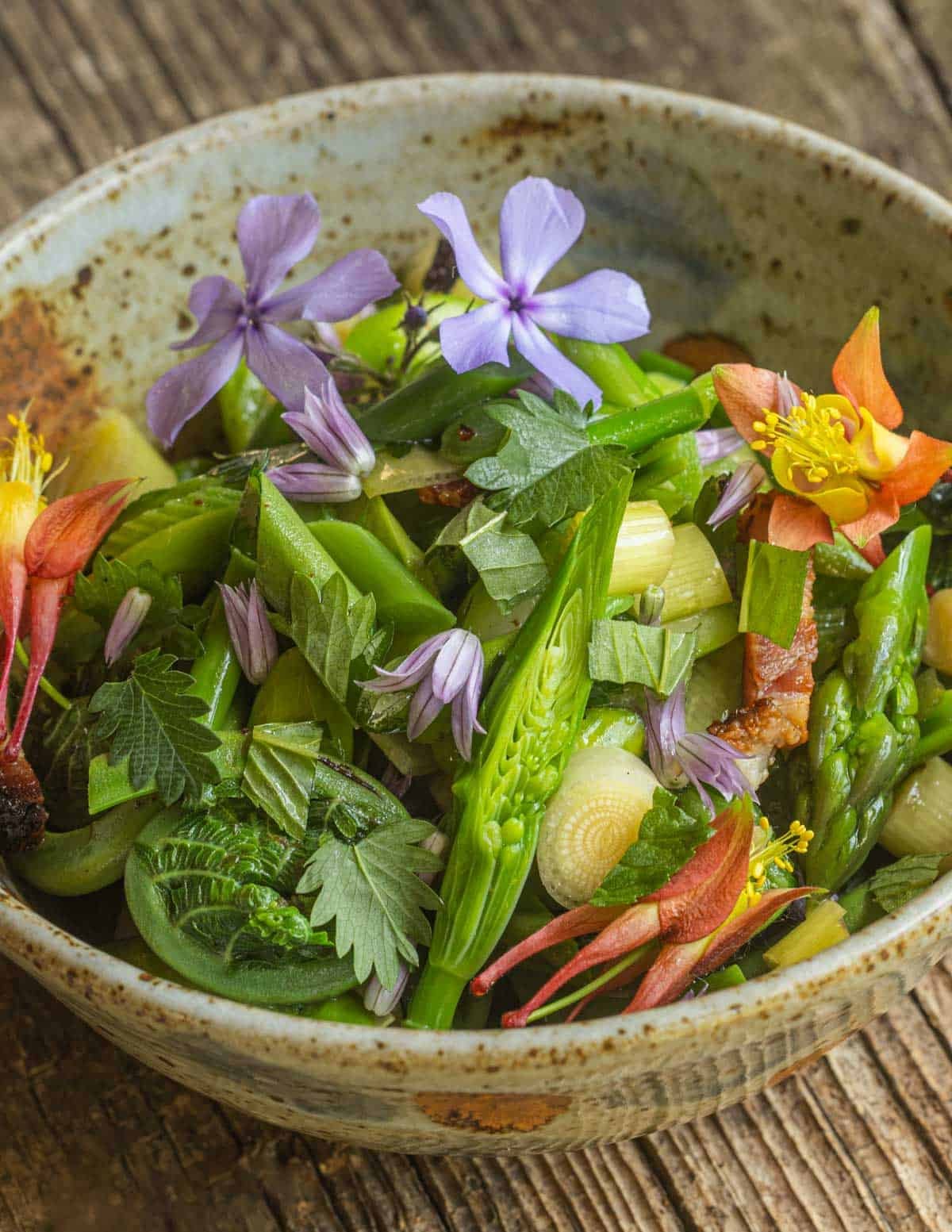 A bowl of shoot vegetables, herbs and flowers garnished with salad burnet leaves and herbs.